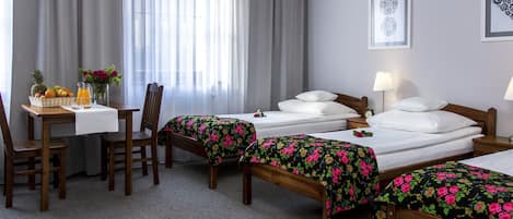 A hotel room with two single beds, a wooden desk with a table, chairs, a vase with flowers, and a fruit basket.