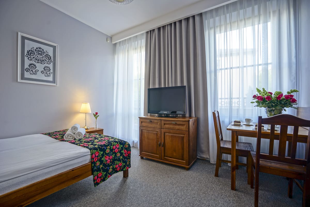 A hotel room with a bed, a TV on a wooden cabinet, a desk with a chair, and a window with curtains.