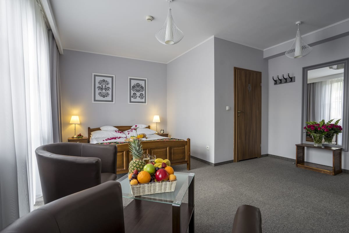 A hotel room with a bed, a dining table with fruit, two chairs, a nightstand, a lamp, a mirror, and a door.