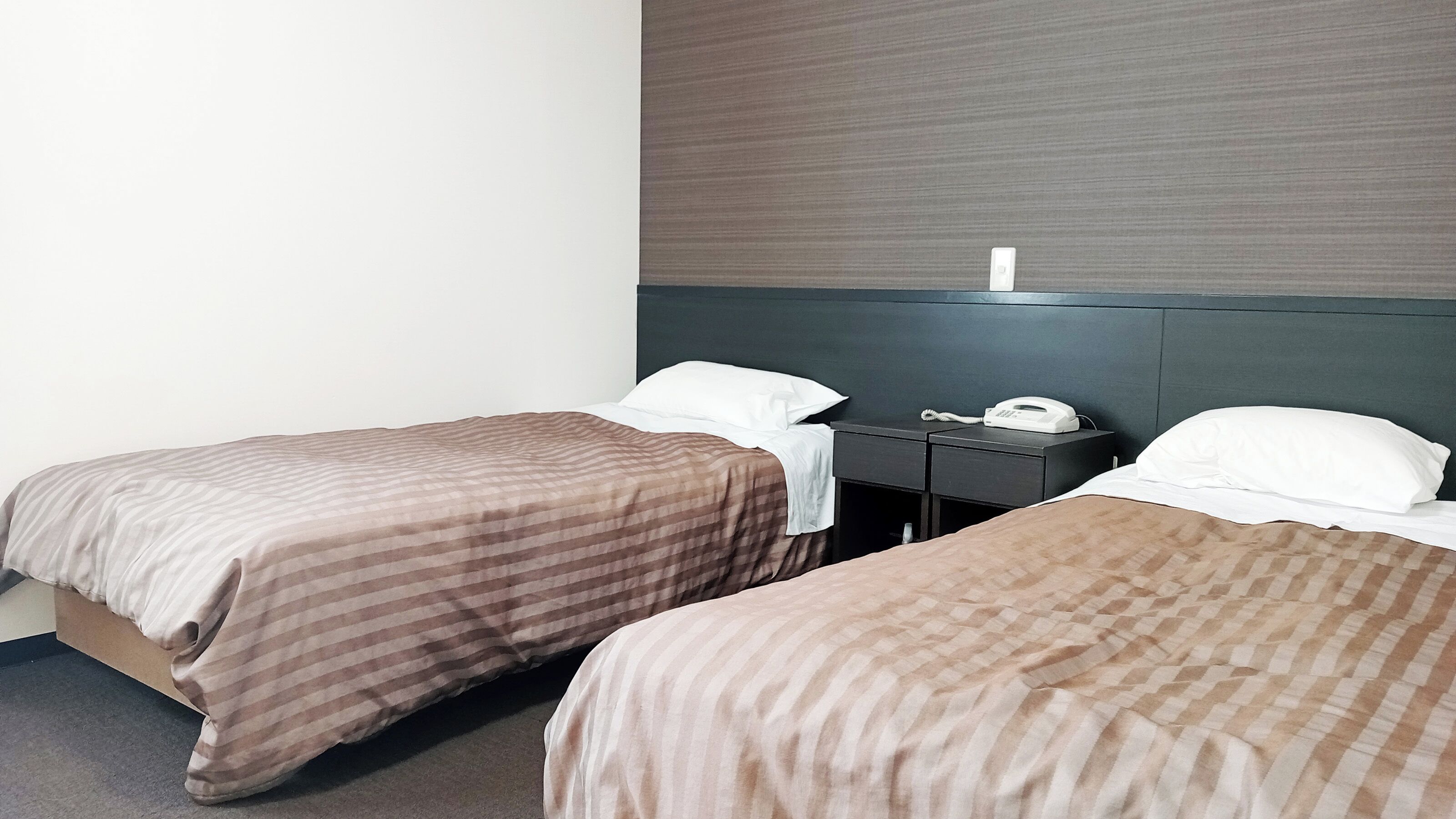 Dos camas individuales con ropa de cama a rayas, una mesita de noche con teléfono y una pared lisa con un panel texturizado.