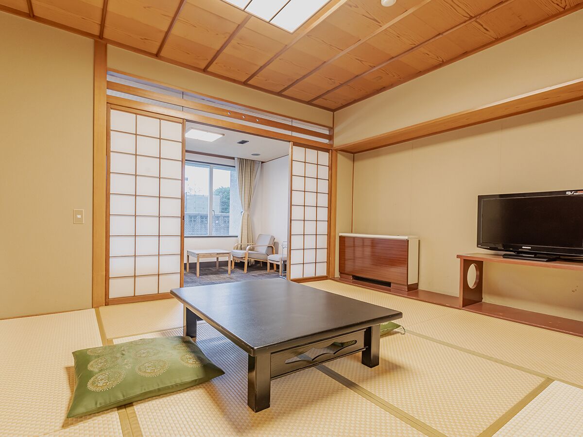 Una habitación con suelo de tatami, una mesa de tatami, un cojín verde, un televisor y una puerta corredera que da a un balcón.