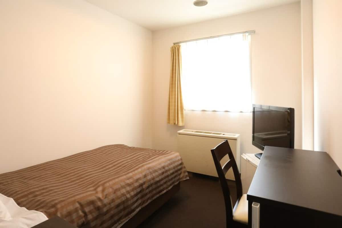 Quarto pequeno com uma cama de solteiro, escrivaninha, cadeira e televisão.