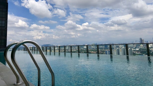 Una piscina infinita con vistas a un horizonte urbano, barandillas metálicas y un cielo parcialmente nublado.