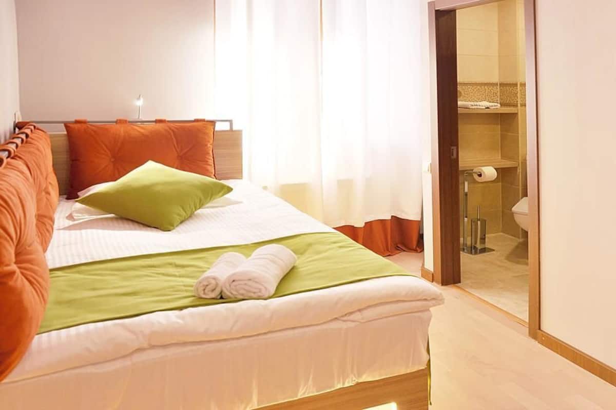 A hotel room with a bed, green and orange pillows, a neatly made bed with towels, a wooden headboard, a bathroom visible through a glass door, and a light-colored floor.