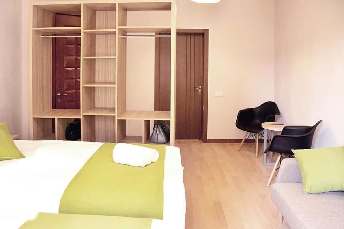 A hotel room with a large wardrobe, a bed with green bedding, a small table with a chair, and a sofa.