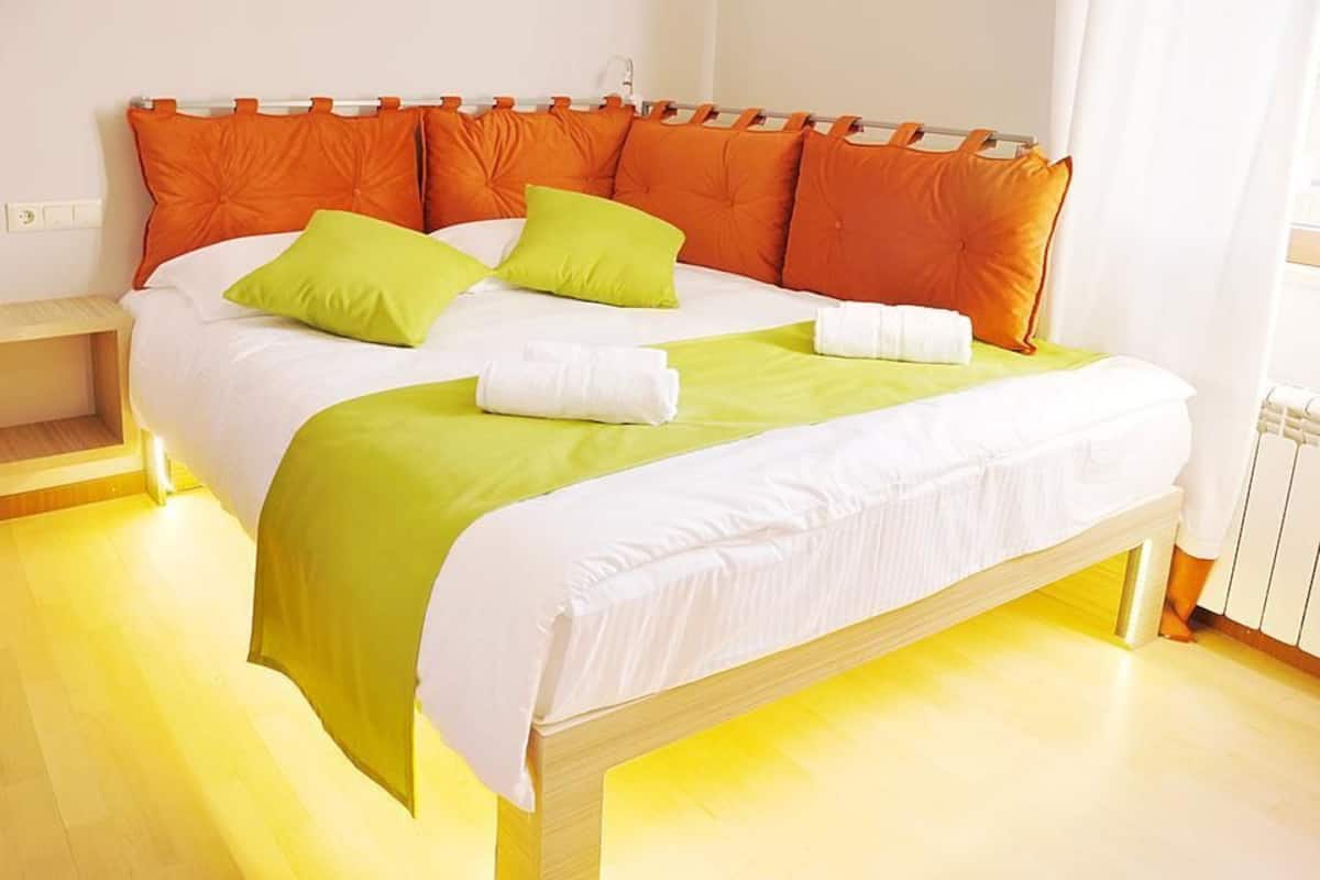 A neatly made bed with a wooden frame, a white mattress, and green and orange pillows.