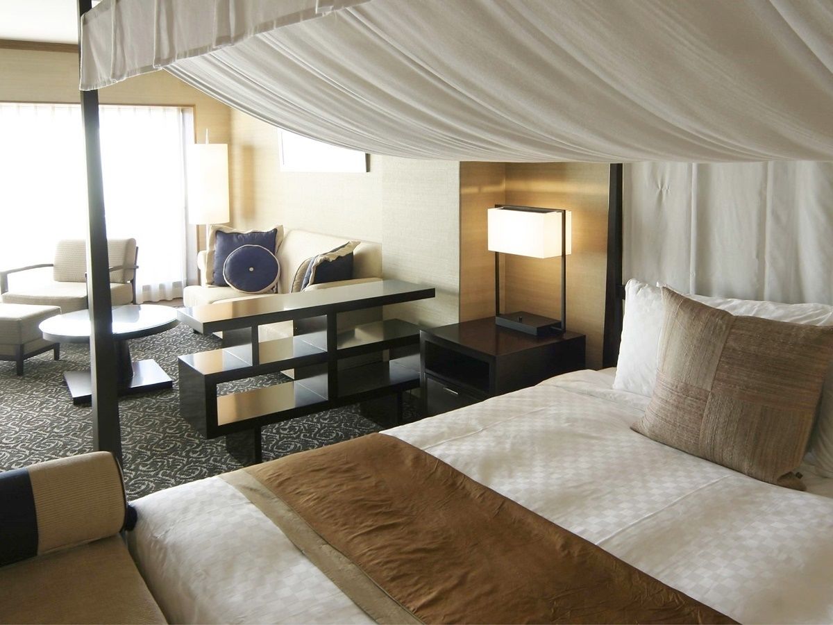 A hotel room with a canopy bed, a desk with a chair, a sofa, and a lamp.