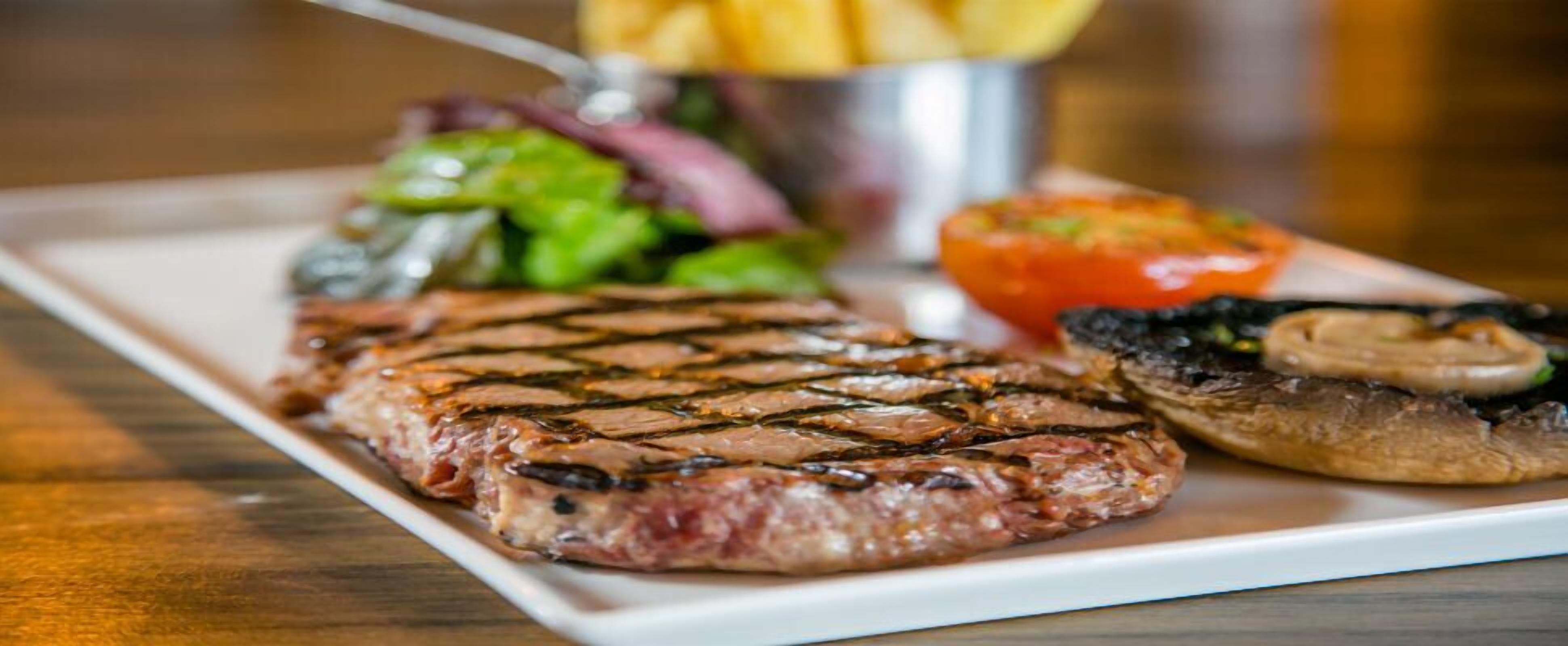Een steak met grillstrepen, een salade, gegrilde champignons en een tomaat op een wit bord.