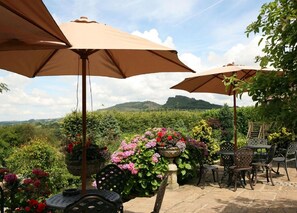 An outdoor seating area with tables and chairs under large umbrellas, surrounded by lush greenery and flowers, with a view of distant hills. - Three Horseshoes Country Inn and Spa (Leek)