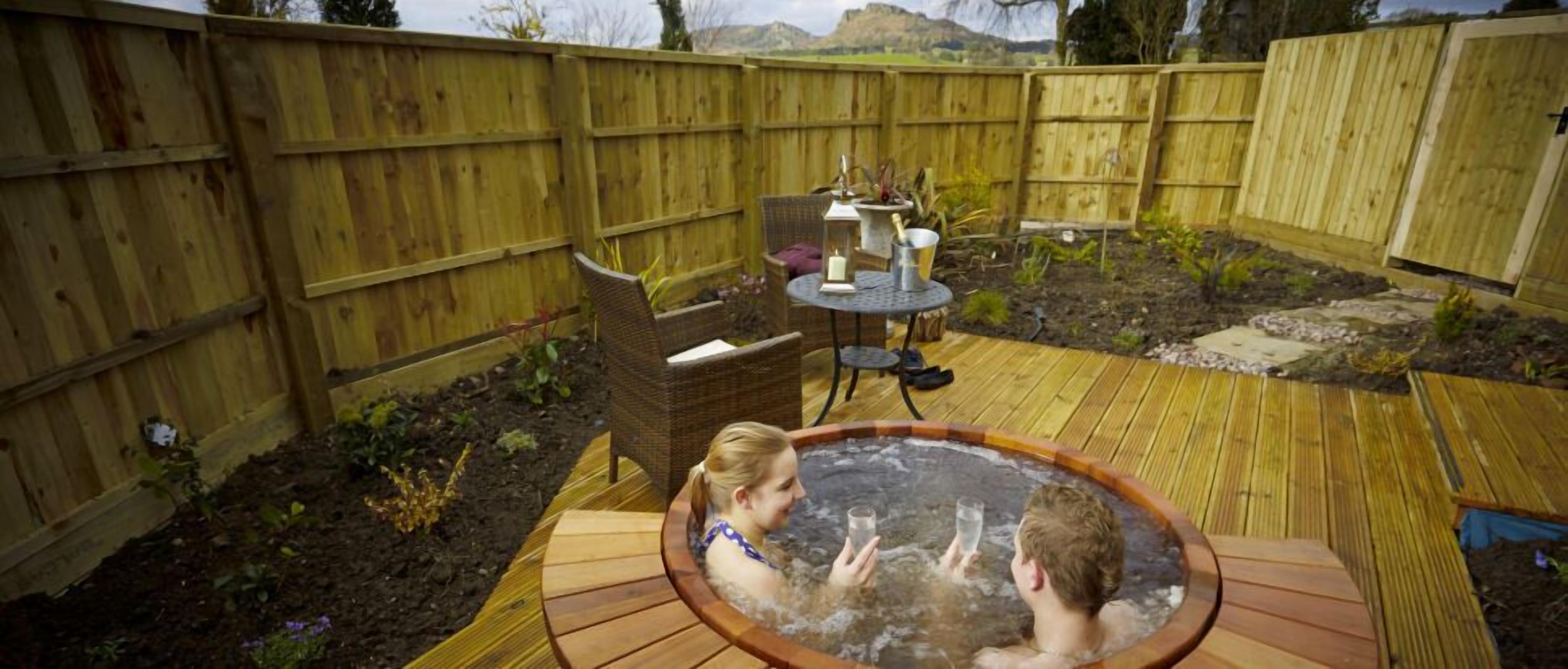 Una vasca idromassaggio in un giardino sul retro con un patio in legno, una sedia in vimini e un tavolo con una candela e un drink.