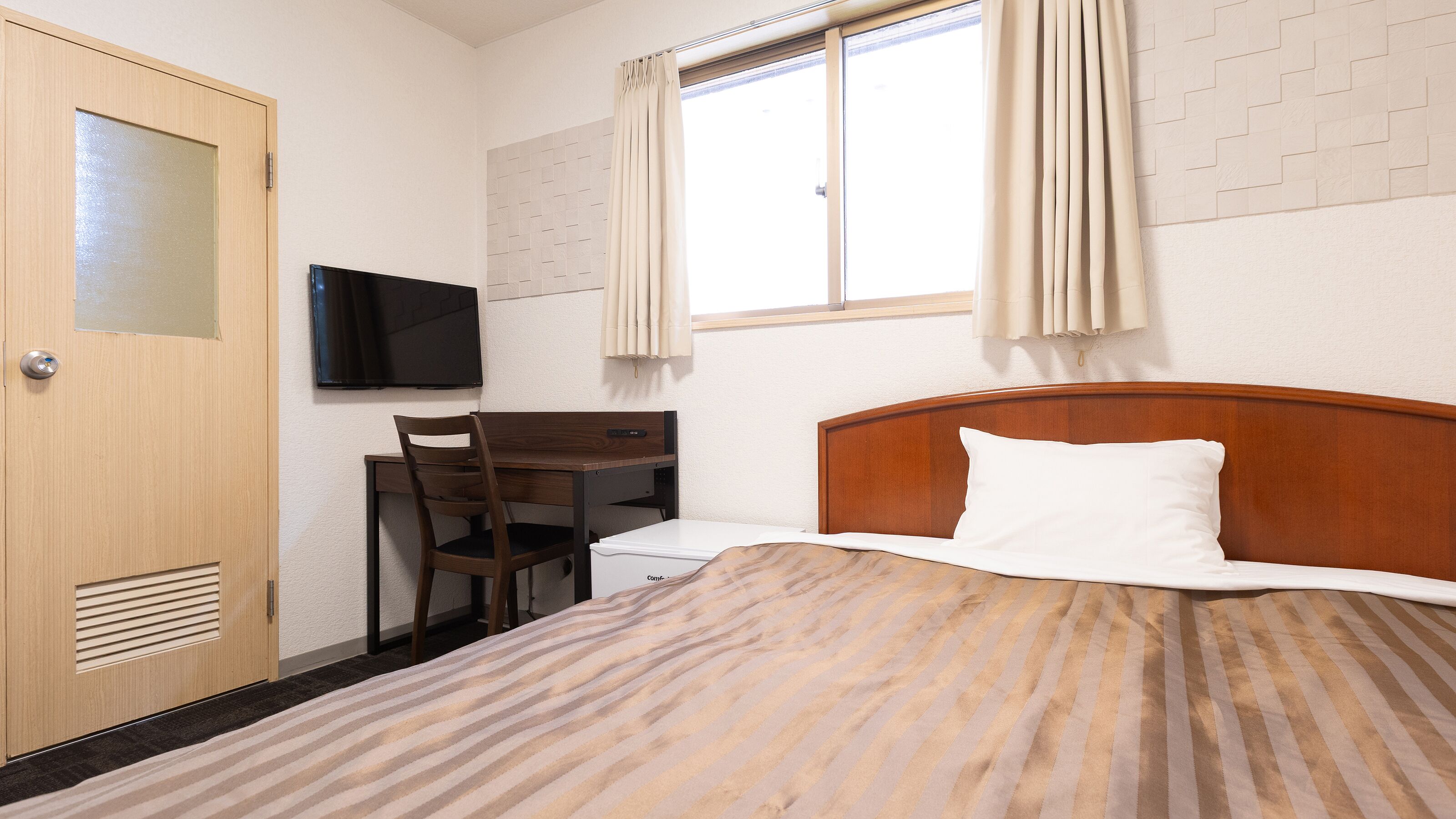 A hotel room with a bed, a desk with a chair, a television, and a window with curtains.