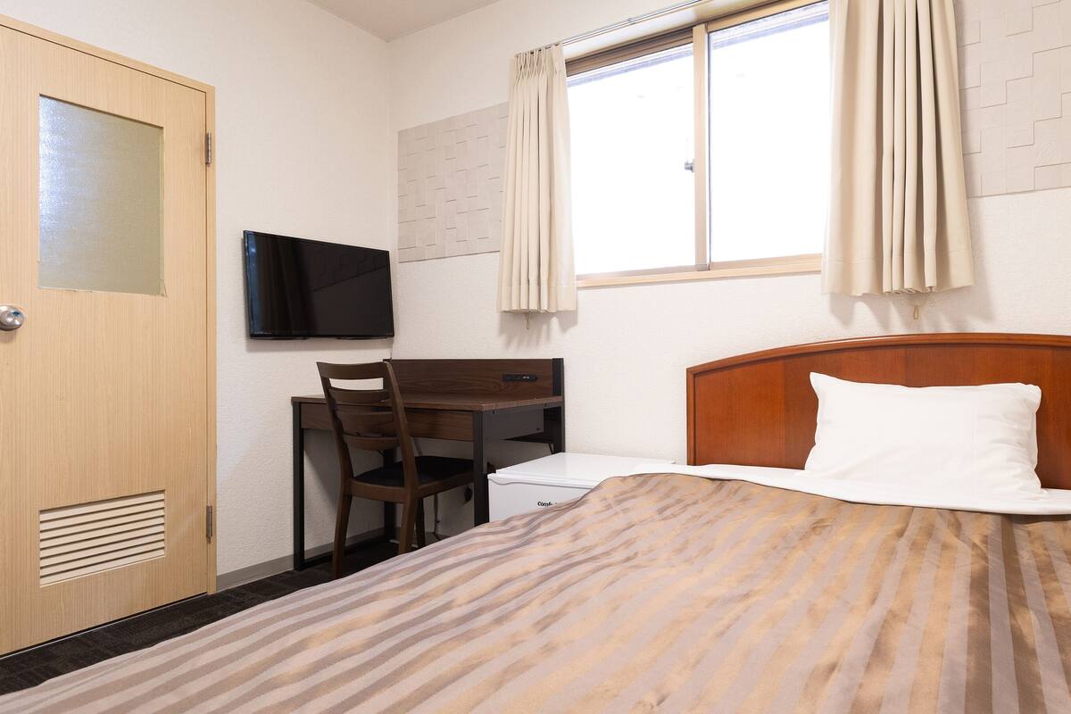 A hotel room with a bed, a desk with a chair, a television, and a window with curtains.