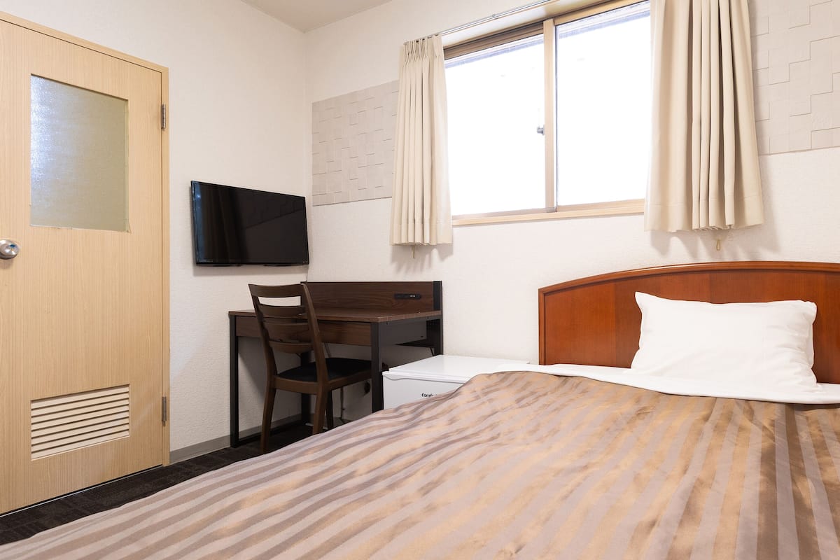 Ein Hotelzimmer mit Bett, Schreibtisch mit Stuhl, Fernseher und einem Fenster mit Vorhängen.