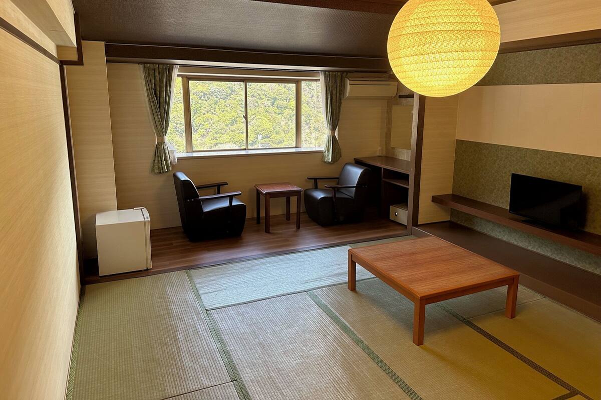 A room with tatami flooring, a low table, two chairs, a television, and a large window with curtains.