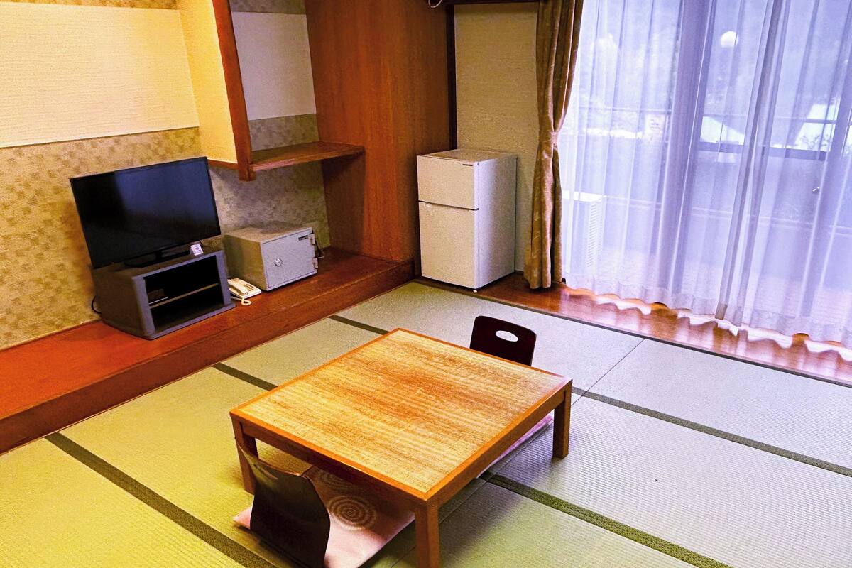 A small room with a wooden table, chairs, a television, a refrigerator, and a window with curtains.