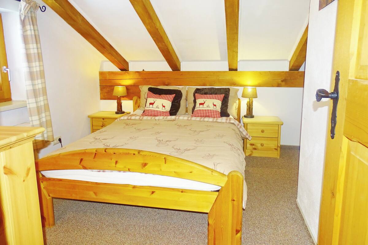 A bedroom with a wooden bed, two bedside tables with lamps, a wooden door, and a window with a curtain.