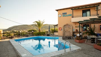 Une piscine avec vue sur les collines, un bâtiment portant l’inscription « SUN VILLAGE », des sièges extérieurs et un anneau de sauvetage.