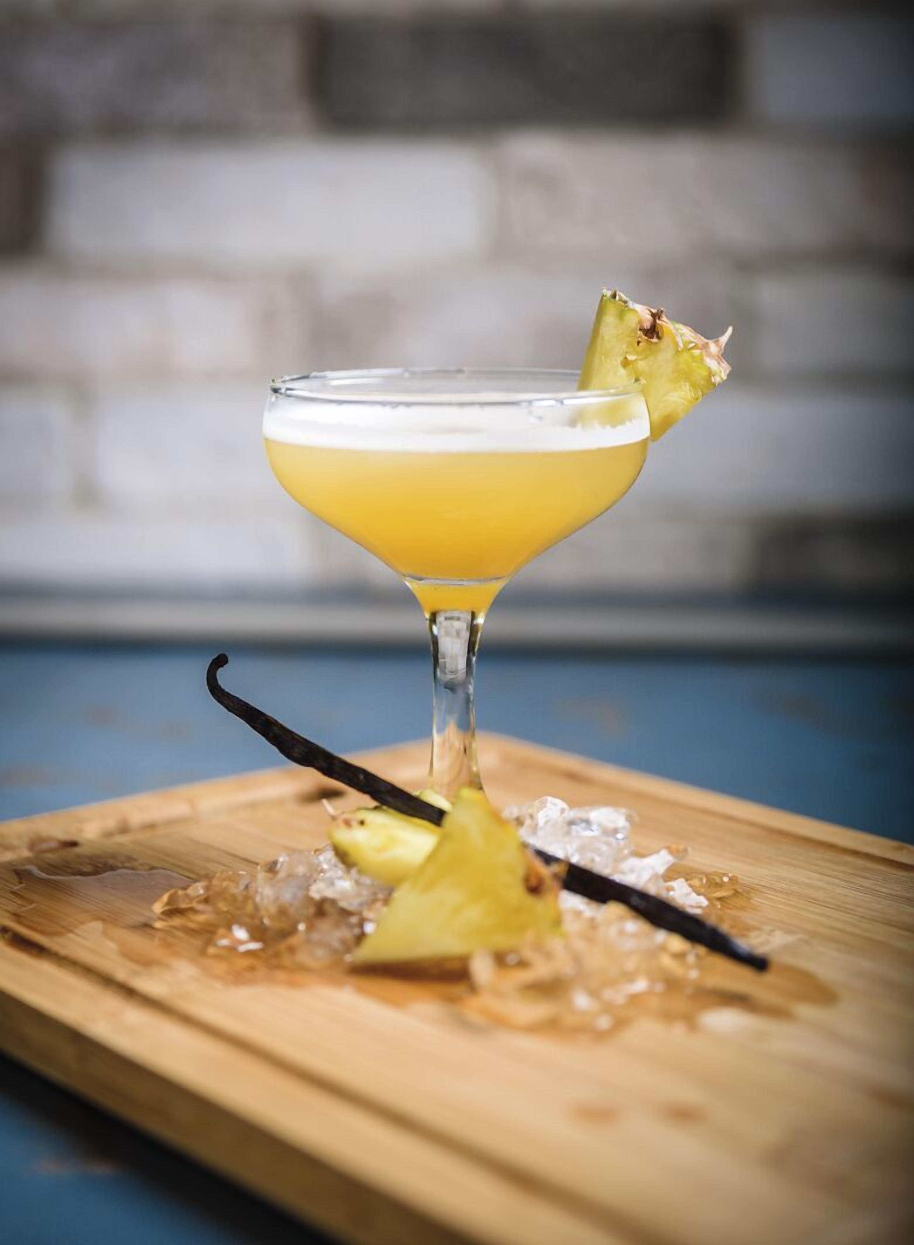Cóctel en copa de martini con una rodaja de piña y una vaina de vainilla sobre una tabla de madera con hielo.