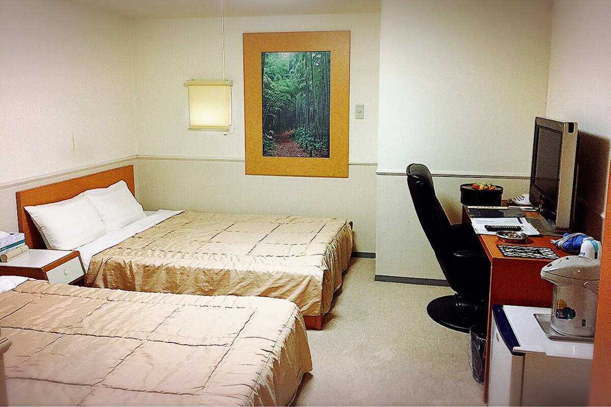 A hotel room with two beds, a desk with a computer and a television, and a painting of a forest on the wall.