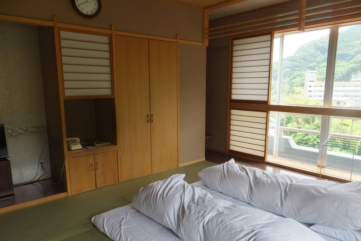 A room with a bed, wooden wardrobe, and a window with a view of buildings and greenery.