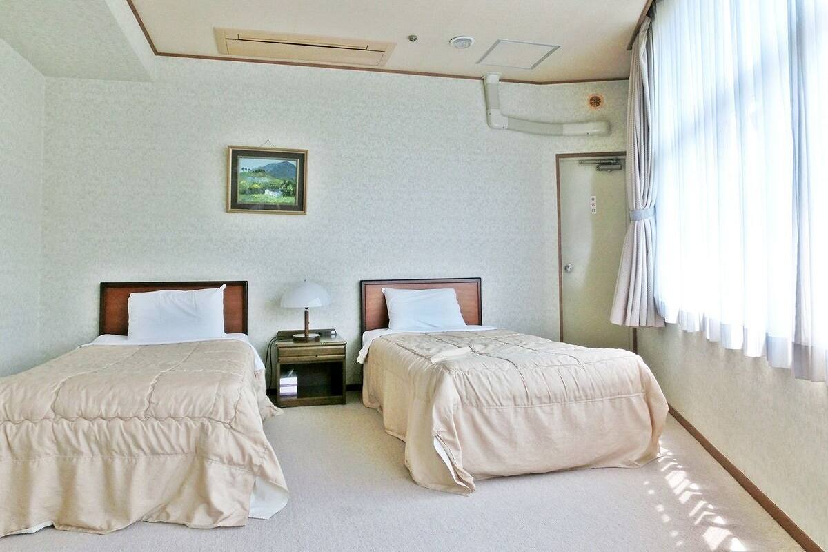A hotel room with two beds, a nightstand, a lamp, a picture on the wall, and a window with curtains.