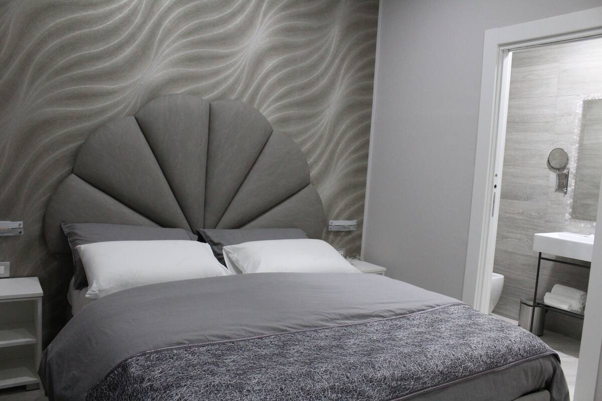 Un dormitorio moderno con un cabecero de diseño original en forma de abanico, una colcha gris y una pared blanca con un patrón ondulado.