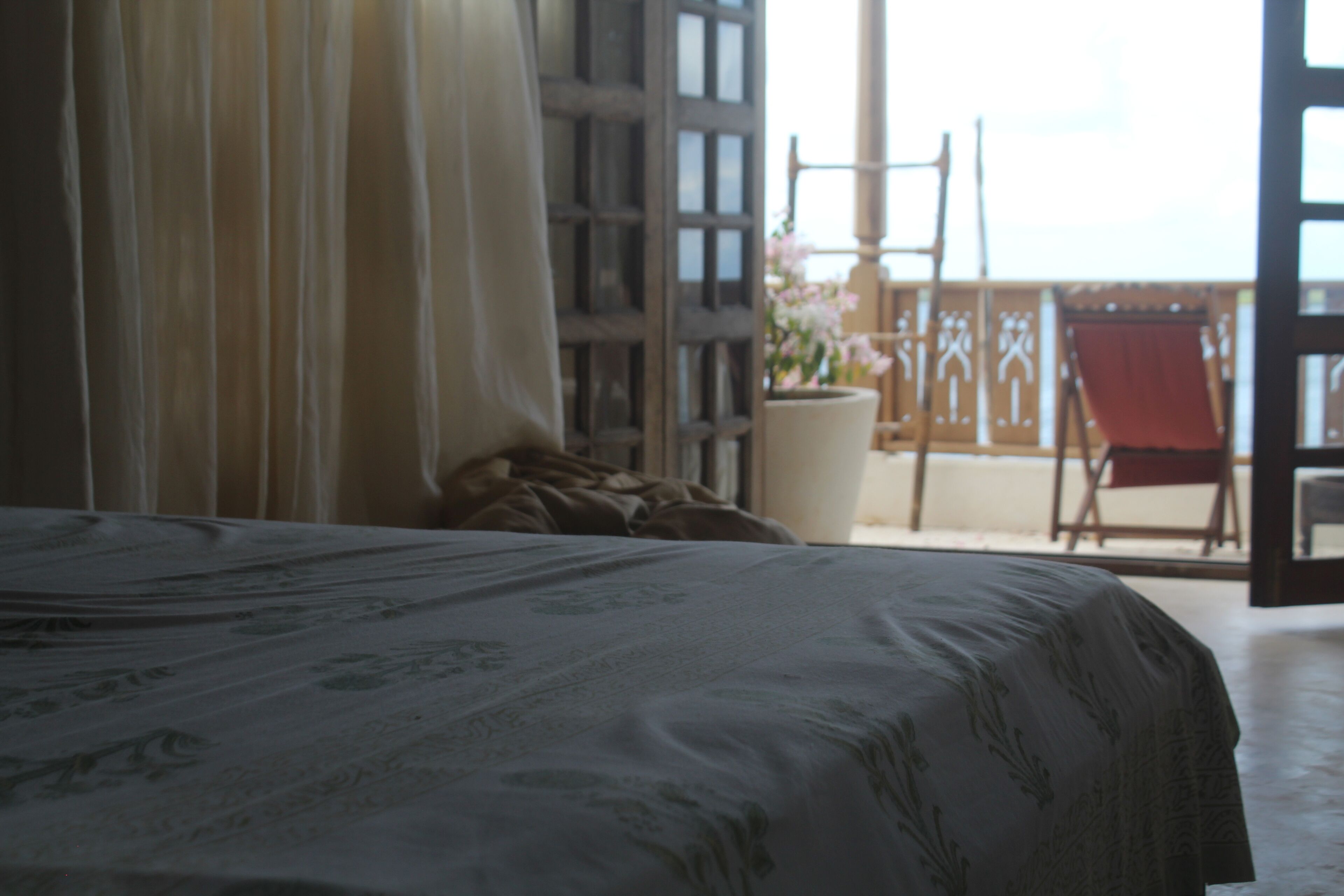 A bedroom with a bed, a window with curtains, a balcony with a chair, and a potted plant.