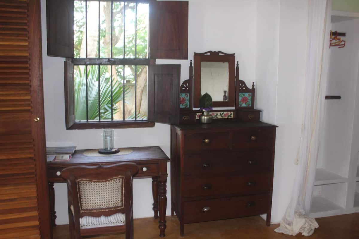 A room with a wooden desk, a chair, a dresser, and a mirror. There is a window with a view of greenery outside.
