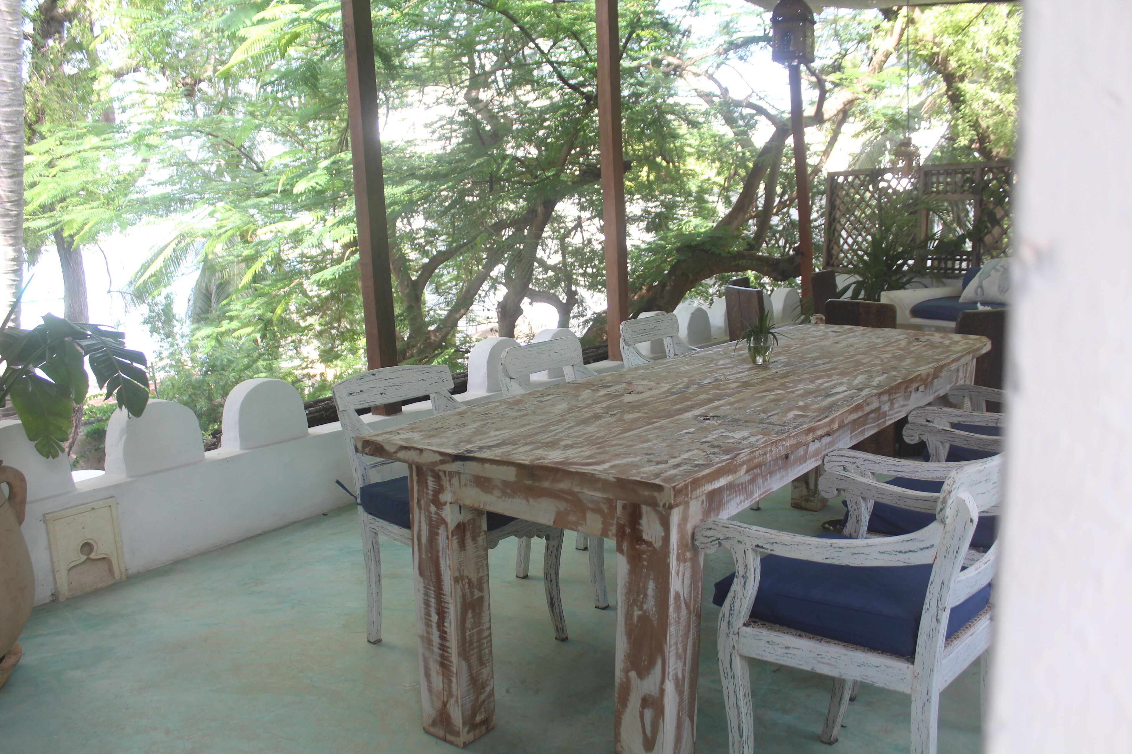 A rustic outdoor dining area with a wooden table and white chairs, surrounded by lush greenery.