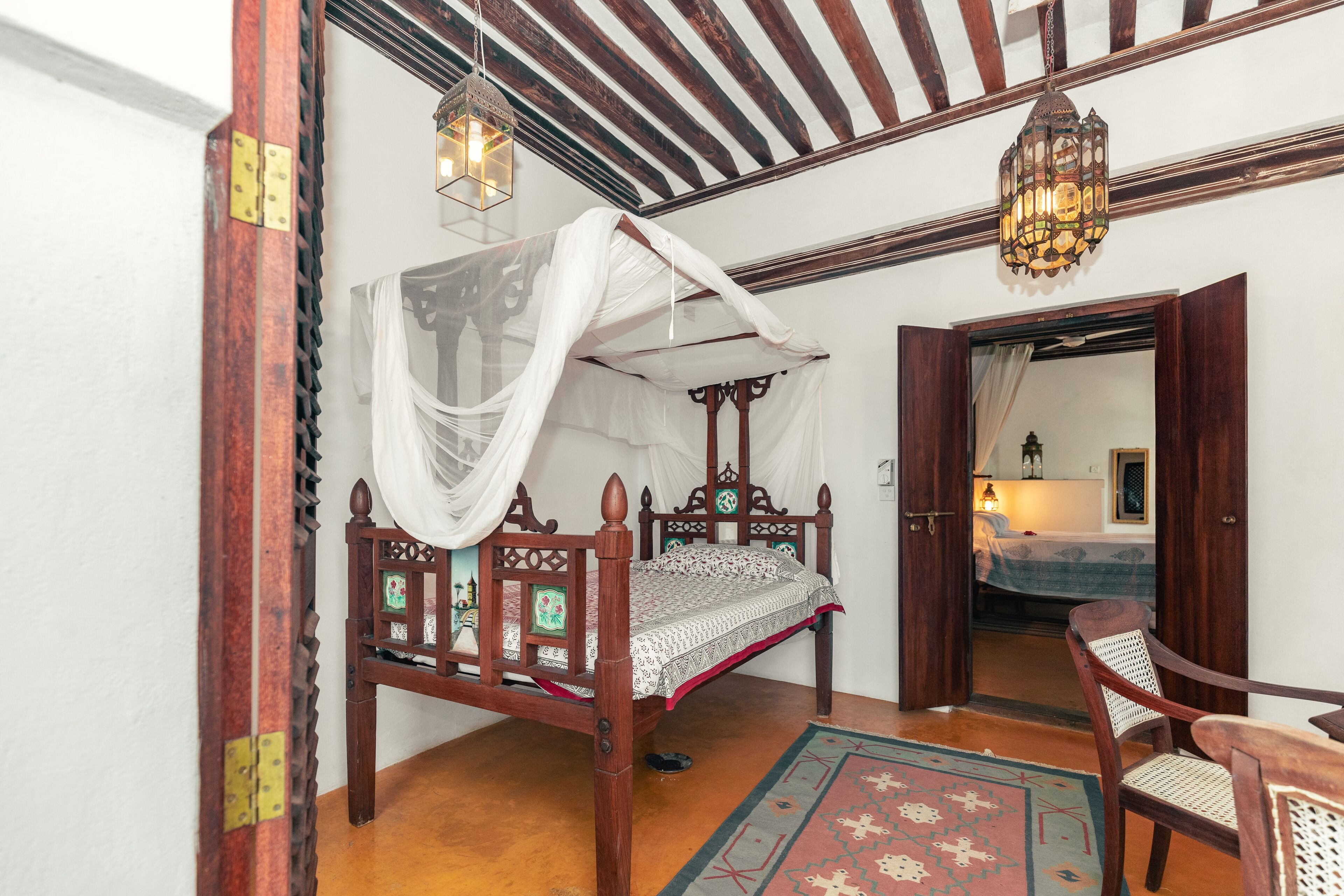 A traditional wooden four-poster bed with a canopy, a wooden chair, and a patterned rug in a room with exposed wooden beams and a hanging lantern.
