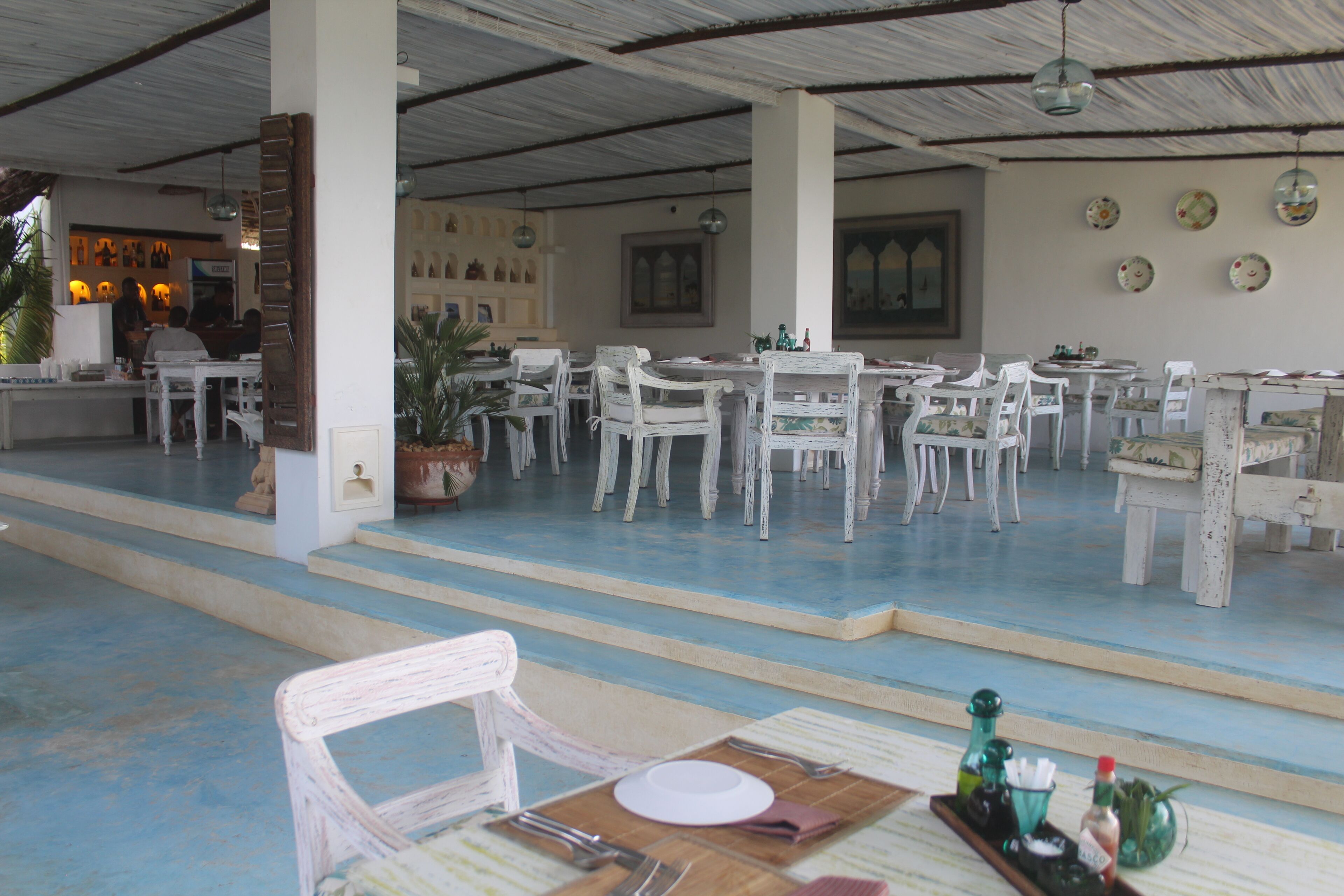 A spacious dining area with white chairs and tables, a bar area in the background, and a decorative wall with plates.