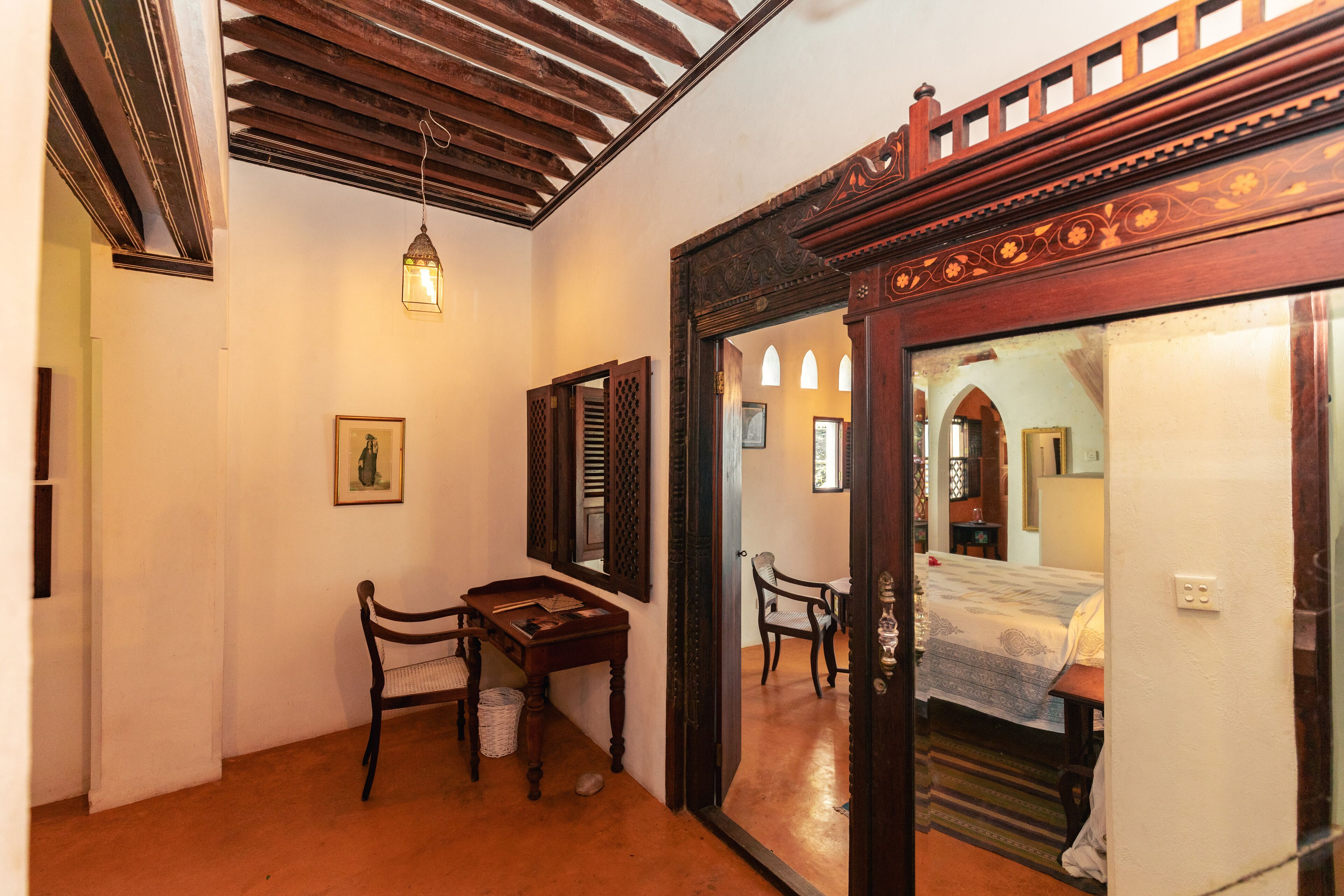 A room with a wooden ceiling, a desk with a chair, a bed with a patterned bedspread, and a mirror with decorative woodwork.