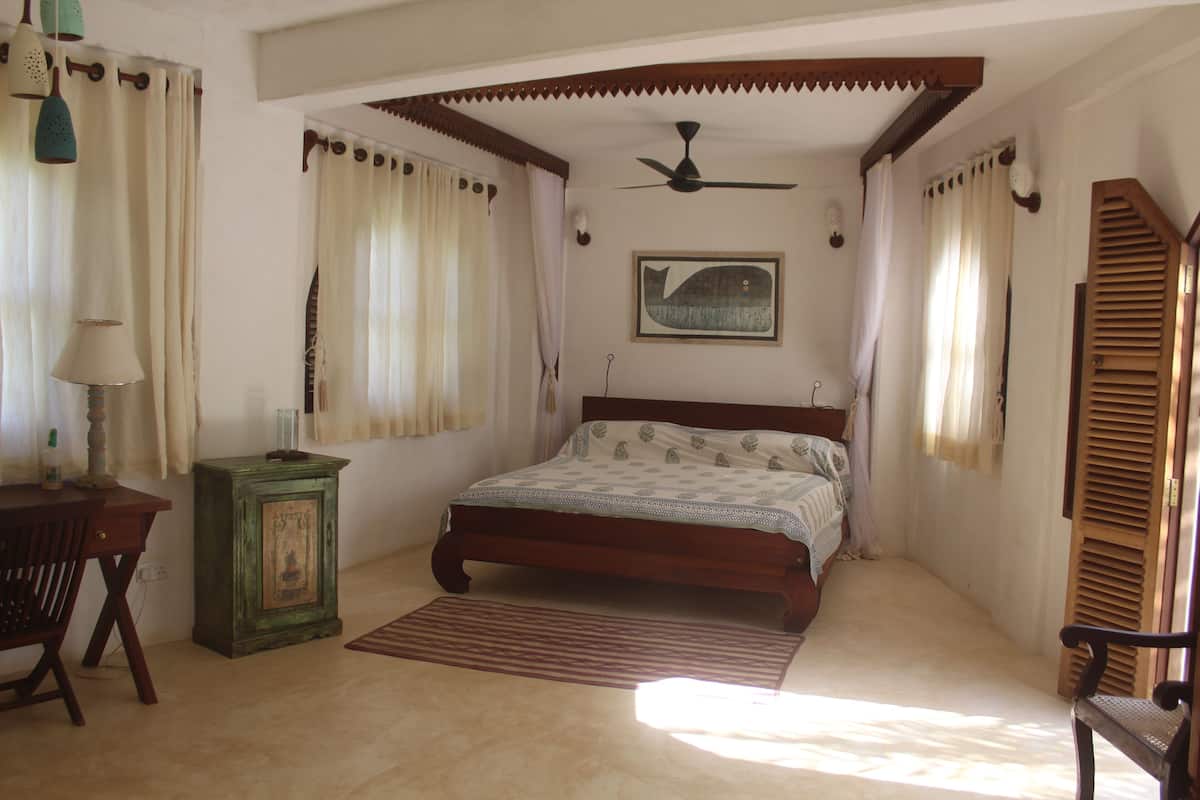 A bedroom with a bed, a wooden headboard, a ceiling fan, a bedside table, a lamp, a green cabinet, and a window with curtains.