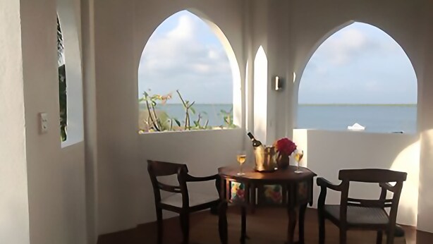 A room with two arched windows overlooking a body of water, a table with chairs, and a bottle of champagne. - Kijani Hotel (Lamu)
