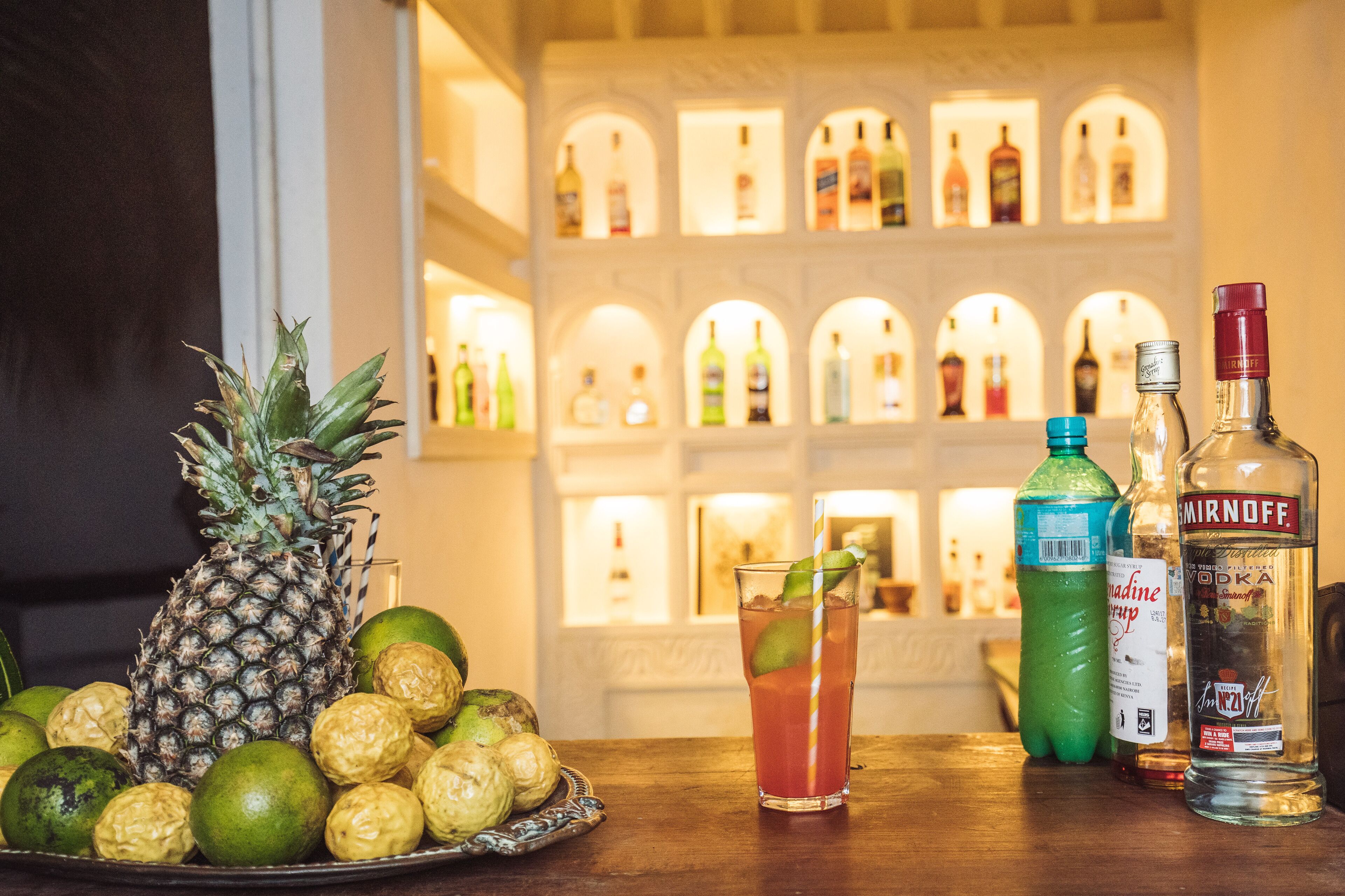 Un bar avec un ananas, des citrons verts et un verre posés sur une table en bois, et en arrière-plan, des bouteilles d’alcool soigneusement alignées.