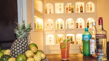 Un bar avec un ananas, des citrons verts et un verre posés sur une table en bois, et en arrière-plan, des bouteilles d’alcool soigneusement alignées.