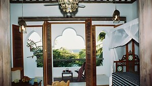 A room with a bed, a wooden chair, a small table, and a window with a view of greenery. - Kijani Hotel (Lamu)