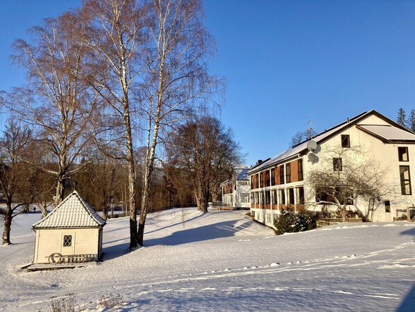 A snow-covered landscape with a small wooden cabin and a large building with multiple windows and a satellite dish. - Sole Gjestegård (Krodsherad)