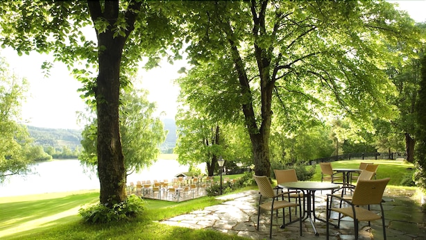 An outdoor patio with stone flooring, surrounded by lush greenery and trees, overlooking a body of water.