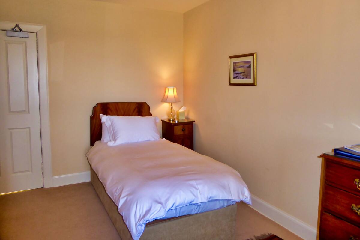 Cama bem arrumada com lençóis brancos, abajur, quadro na parede e cômoda de madeira com gavetas.