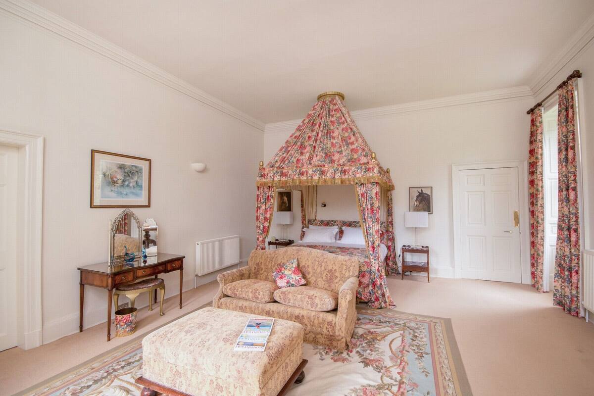 Quarto com cama de dossel, área de estar com poltrona e banquinho, cômoda e um quadro na parede.