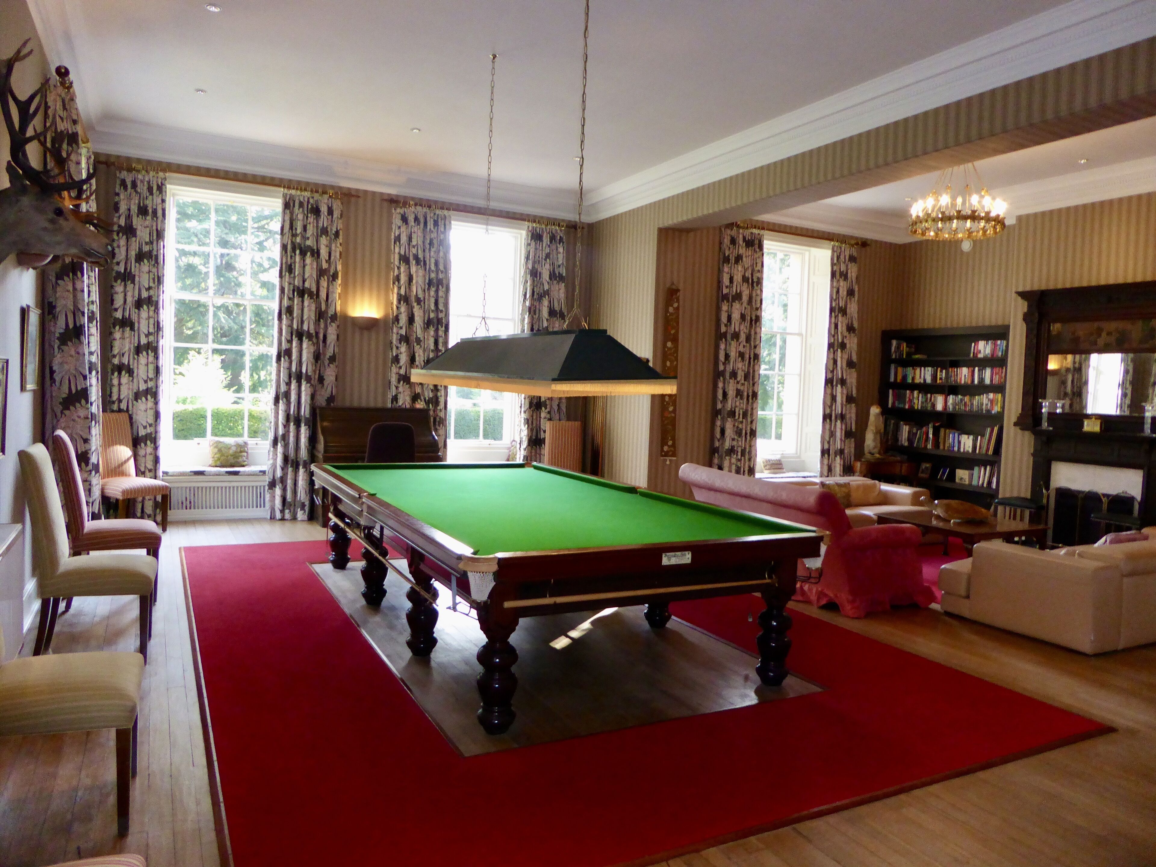 A billiards table in a room with a red carpet, a fireplace, a bookshelf, and a large window with curtains.