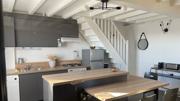 A modern kitchen with a central island, a dining area with chairs, a staircase, and a small seating area with a table and chairs. - Chez Juliette Maison Gîte (Rontignon)