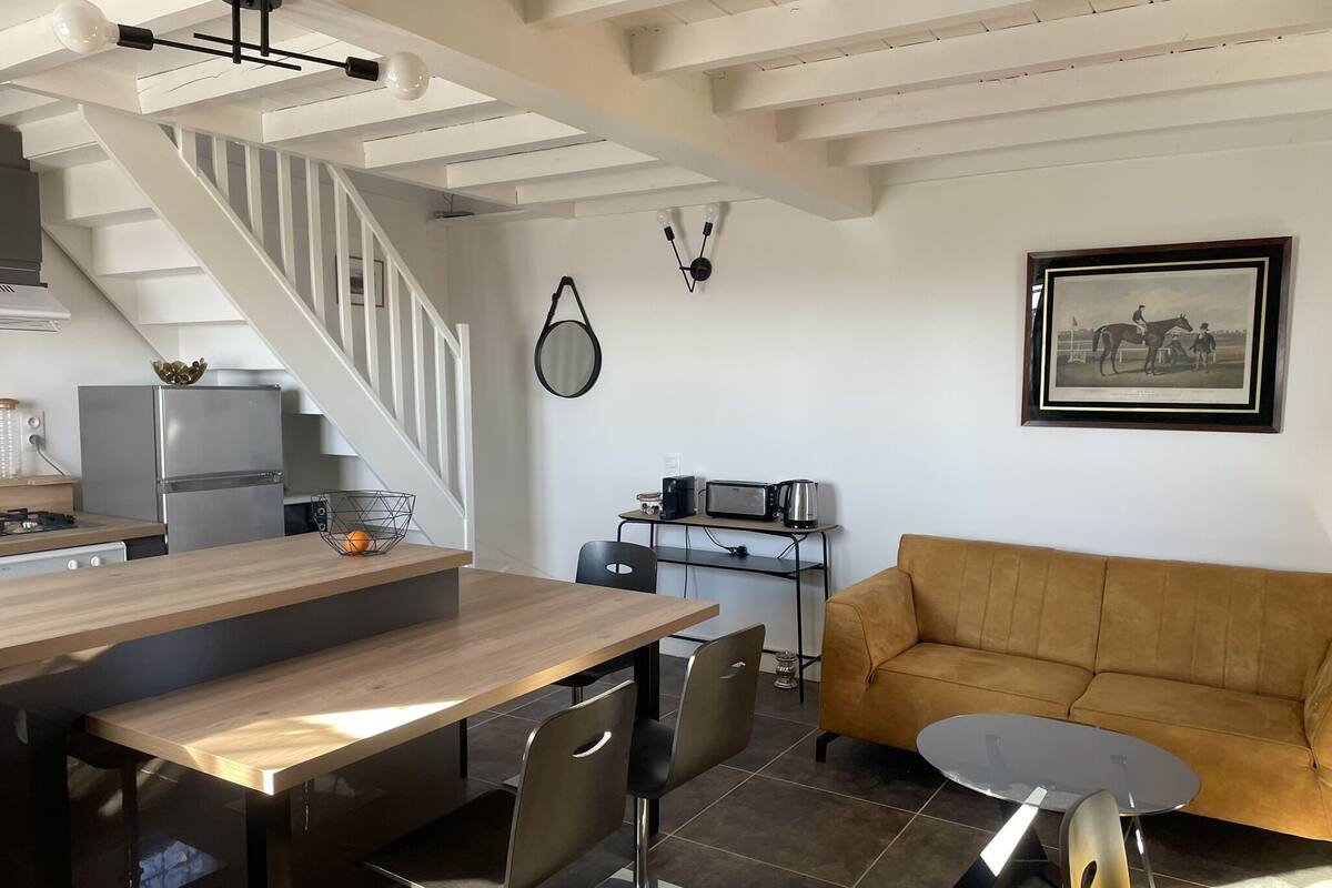 A modern kitchen with a wooden table and chairs, a sofa, and a dining table with chairs. There is a staircase leading to another level.