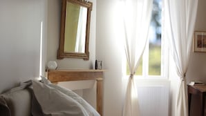 A bedroom with a bed, a bedside table, a mirror, and a window with curtains. - Chez Juliette Maison Gîte (Rontignon)