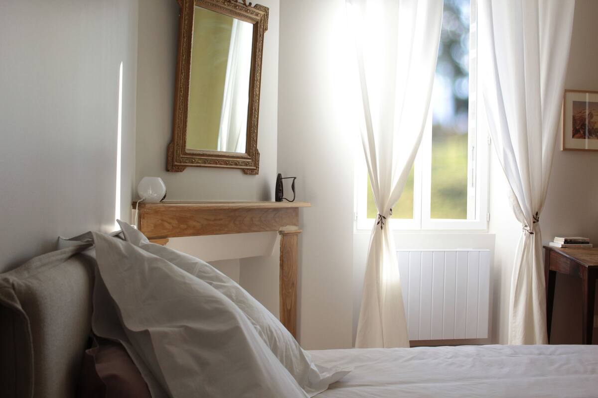 A bedroom with a bed, a bedside table, a mirror, and a window with curtains.
