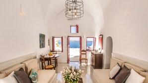 A spacious living area with a high ceiling, a dining table with chairs, a sofa set, and a view of the sea through an open doorway. - Hector Cave House (Santorini)
