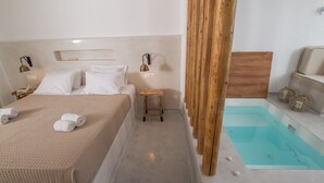A bedroom with a bed, bedside tables, a wooden stool, a small table, and a built-in bathtub. - Hector Cave House (Santorini)
