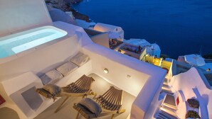 A modern outdoor seating area with a hot tub, lounge chairs, and a view of the sea and coastline. - Hector Cave House (Santorini)