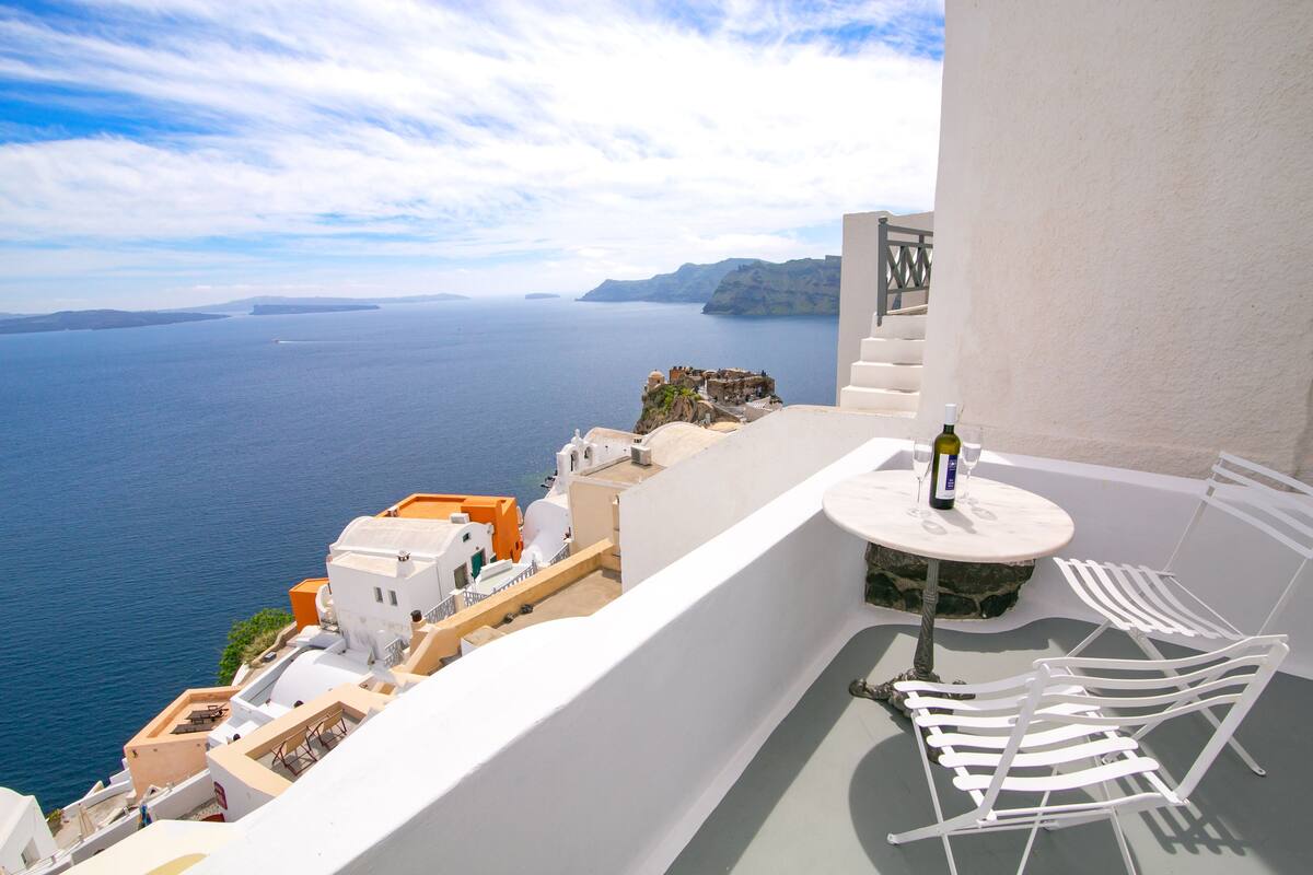 Un balcon avec des chaises blanches et une table, d’où l’on peut admirer une vue sur la côte, avec des bâtiments et la mer.